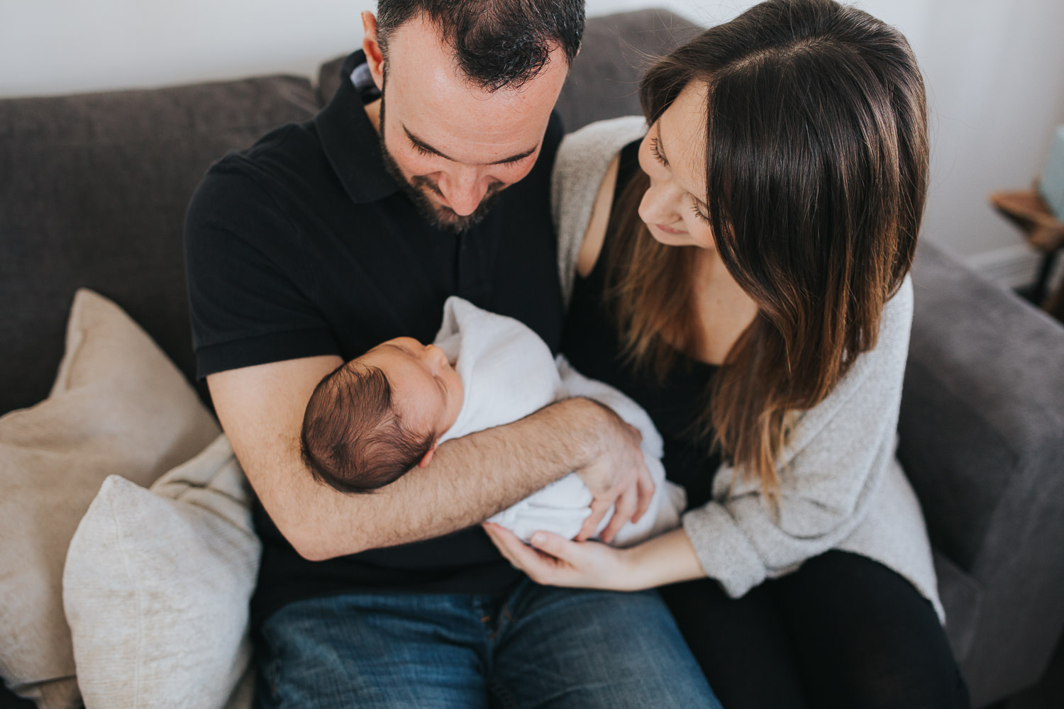 new parents sitting on couch holding and looking at 2 week old baby boy -  Newmarket in-home photos