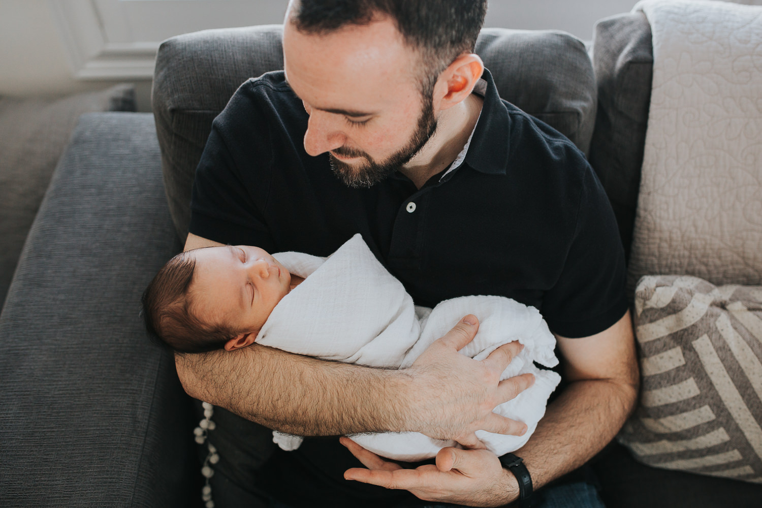 new dad sitting on couch holding and looking at sleeping 2 week old baby son -  Markham in-home photos