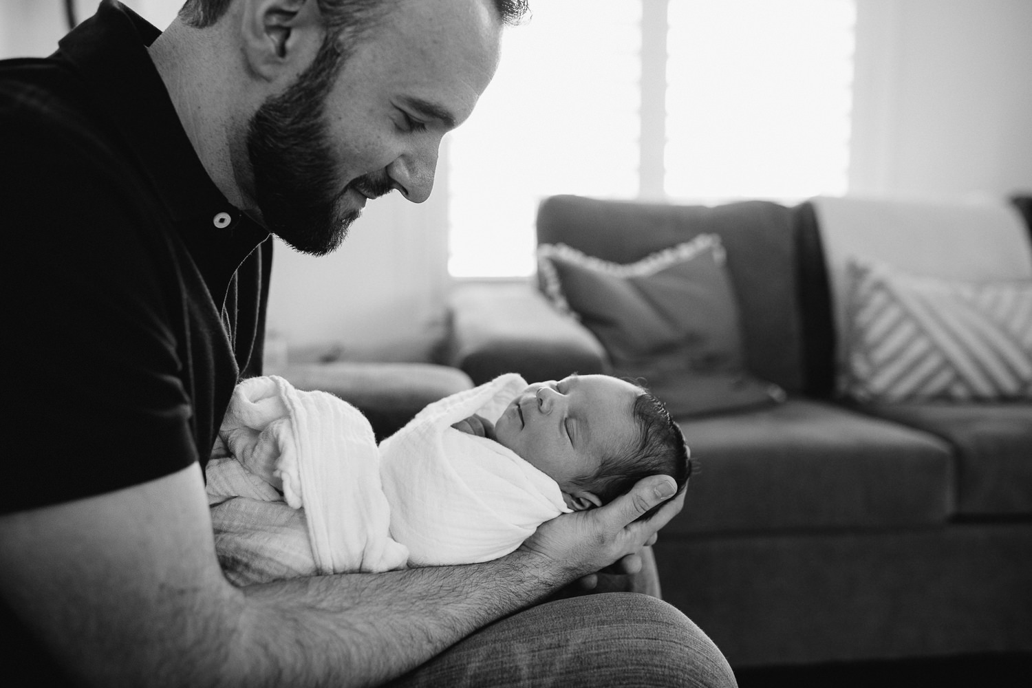 new dad sitting on couch holding and looking at sleeping 2 week old baby boy -  Barrie in-home photos