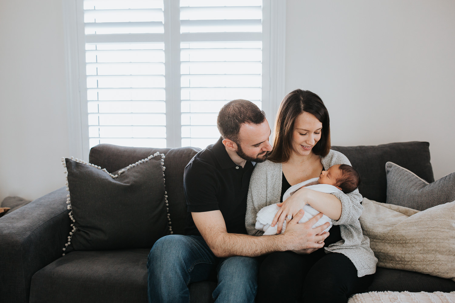 new parents sitting on couch holding and looking at 2 week old baby boy -  Uxbridge in-home photos