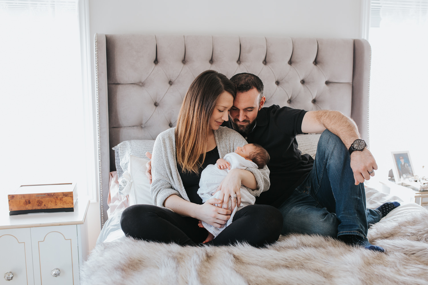 mom and dad sit on master bedroom bed looking at and holding 2 week old baby son - Barrie lifestyle photography