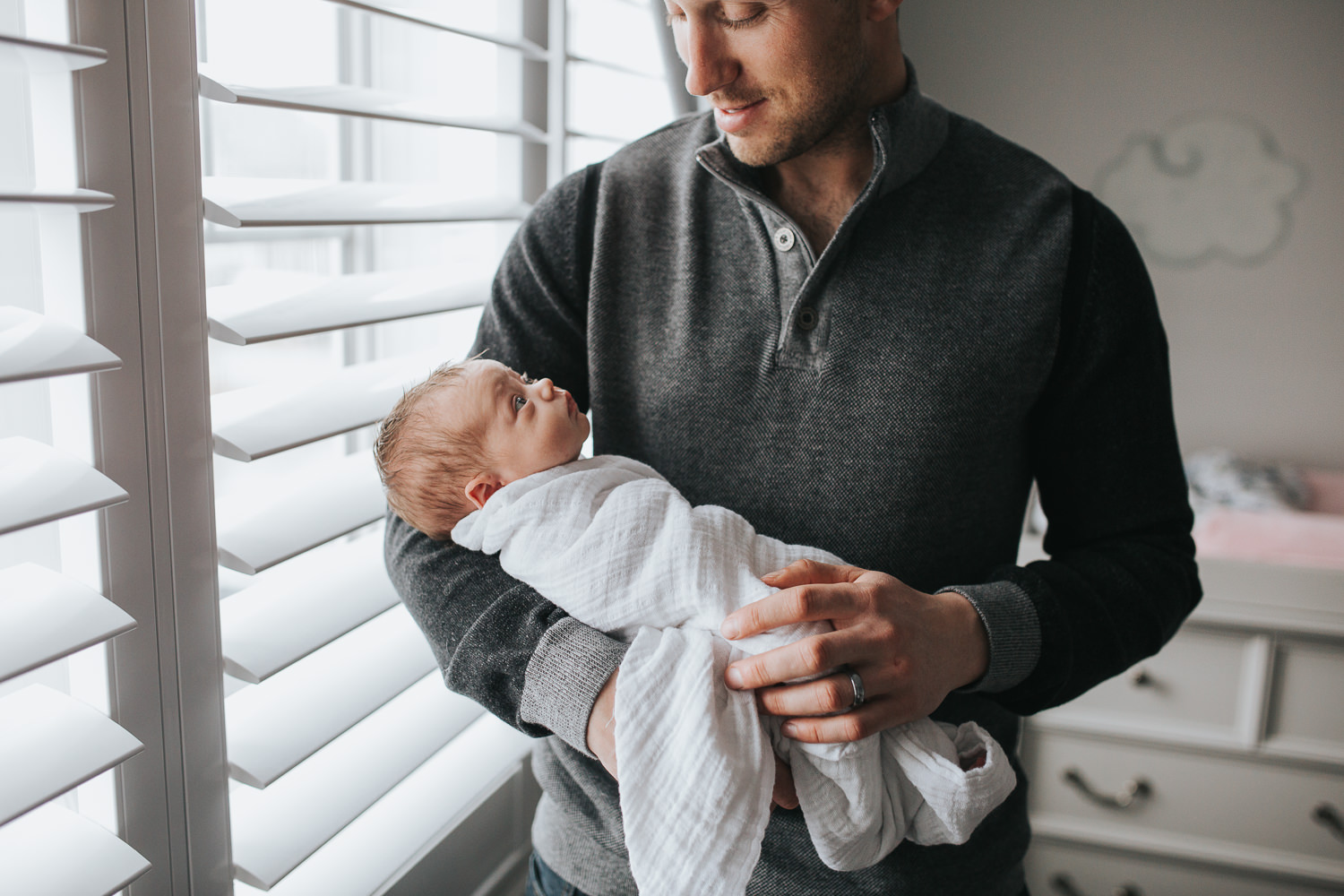 first time father standing in front of nursery window holding 2 week old baby girl, who is awake and looking back at dad- Barrie Lifestyle Photography