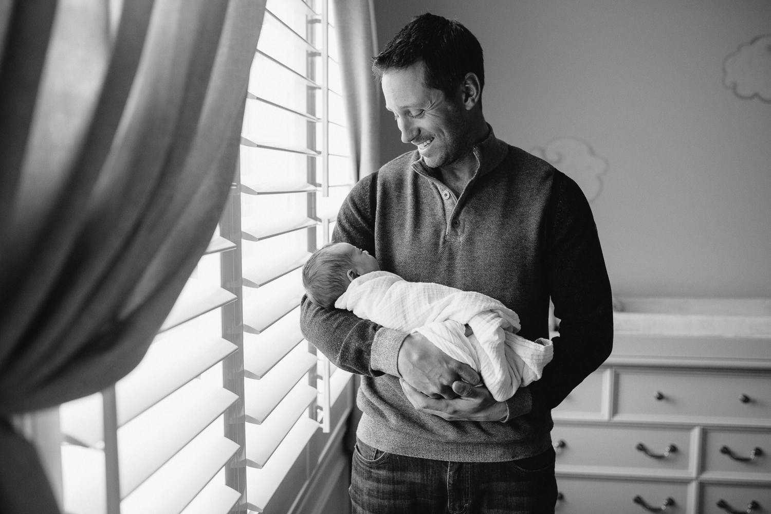 new dad standing in front of nursery window holding and looking at 2 week old baby daughter - Uxbridge Lifestyle Photography