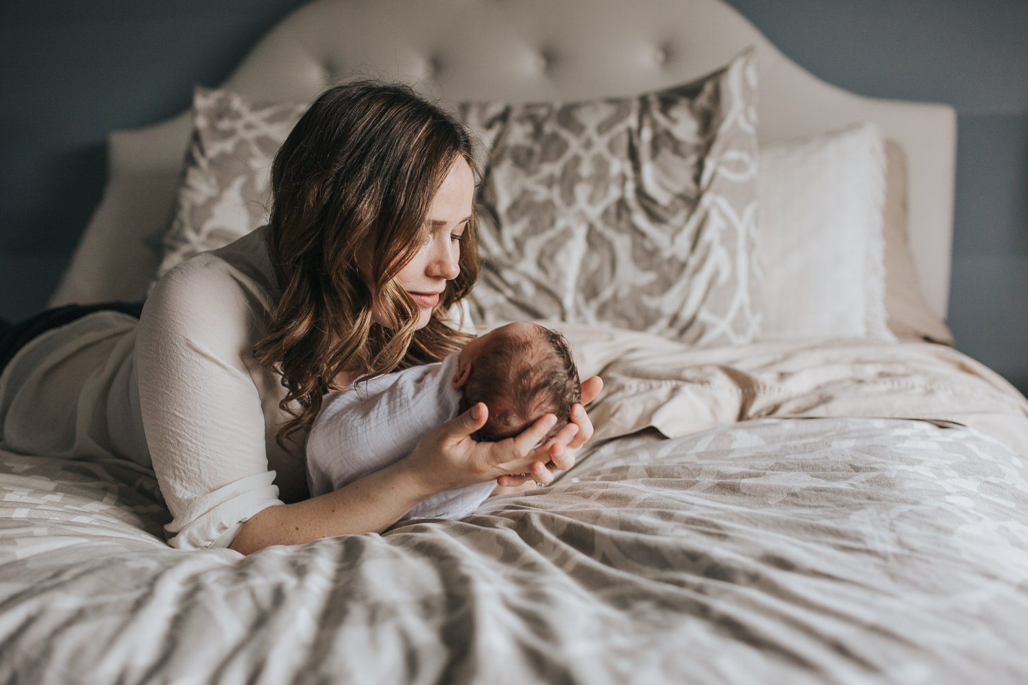 first time mom lying on master bed holding 2 week old baby girl's head in her hands and gazing at her - Stouffville Lifestyle Photos