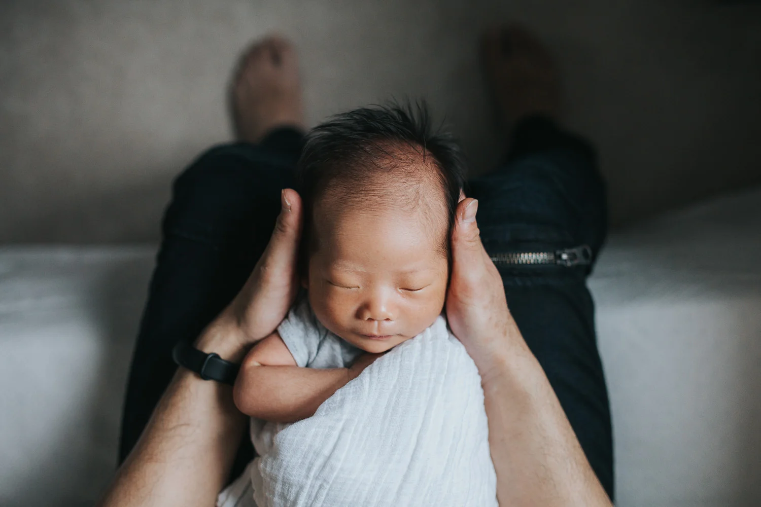 shot from above of sleeping, swaddled 2 week old baby boy lying with head in father's hands - Barrie Lifestyle Photos