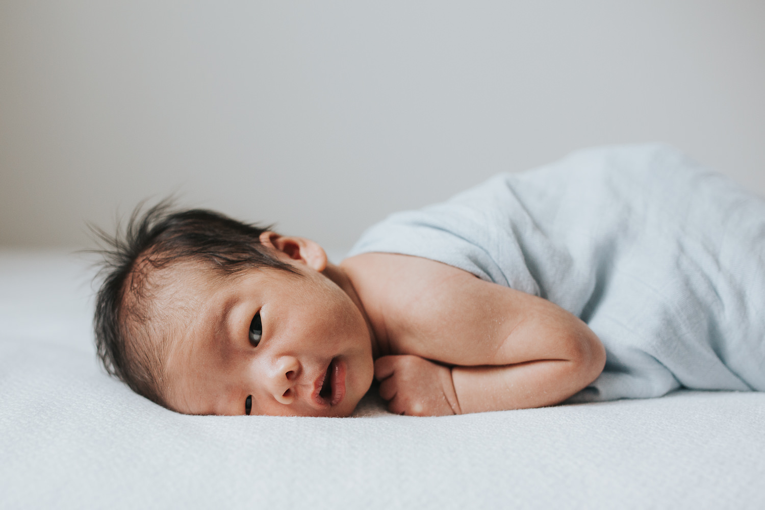 2 week old baby boy in blue swaddle lying on stomach, awake and looking at camera - Stouffville Lifestyle Photography
