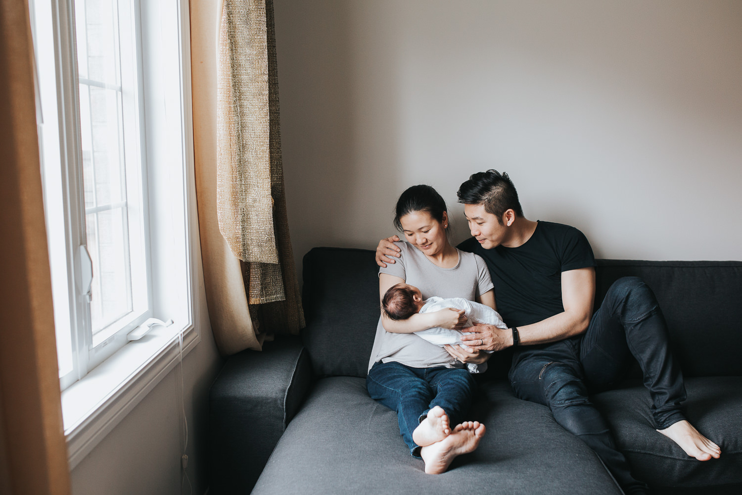 first time parents sitting on couch cuddling and looking at 2 week old baby boy - Stouffville In-Home Photographyf