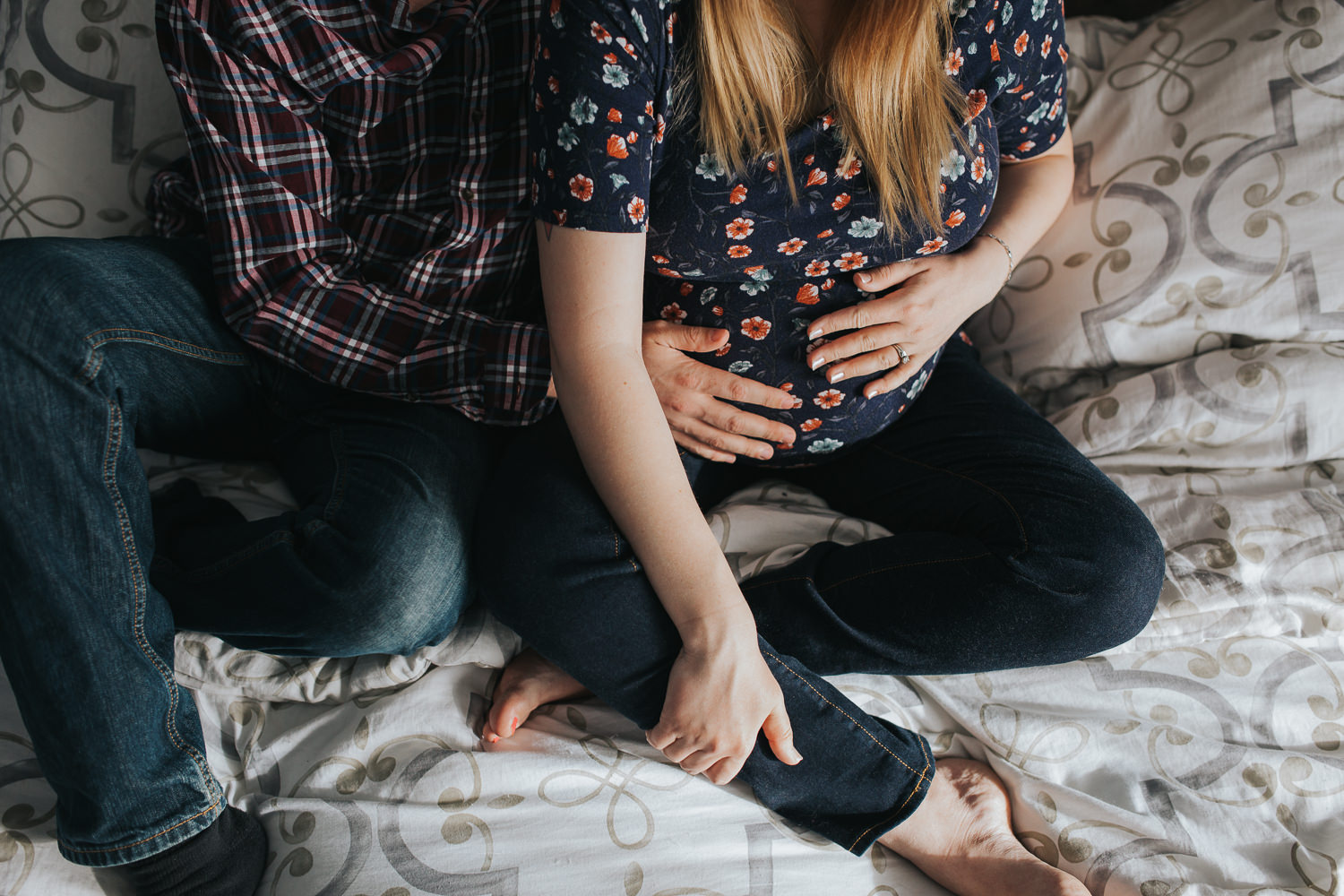 expecting couple snuggles on bed, hands holding wife's pregnant belly - Newmarket In-Home Photography