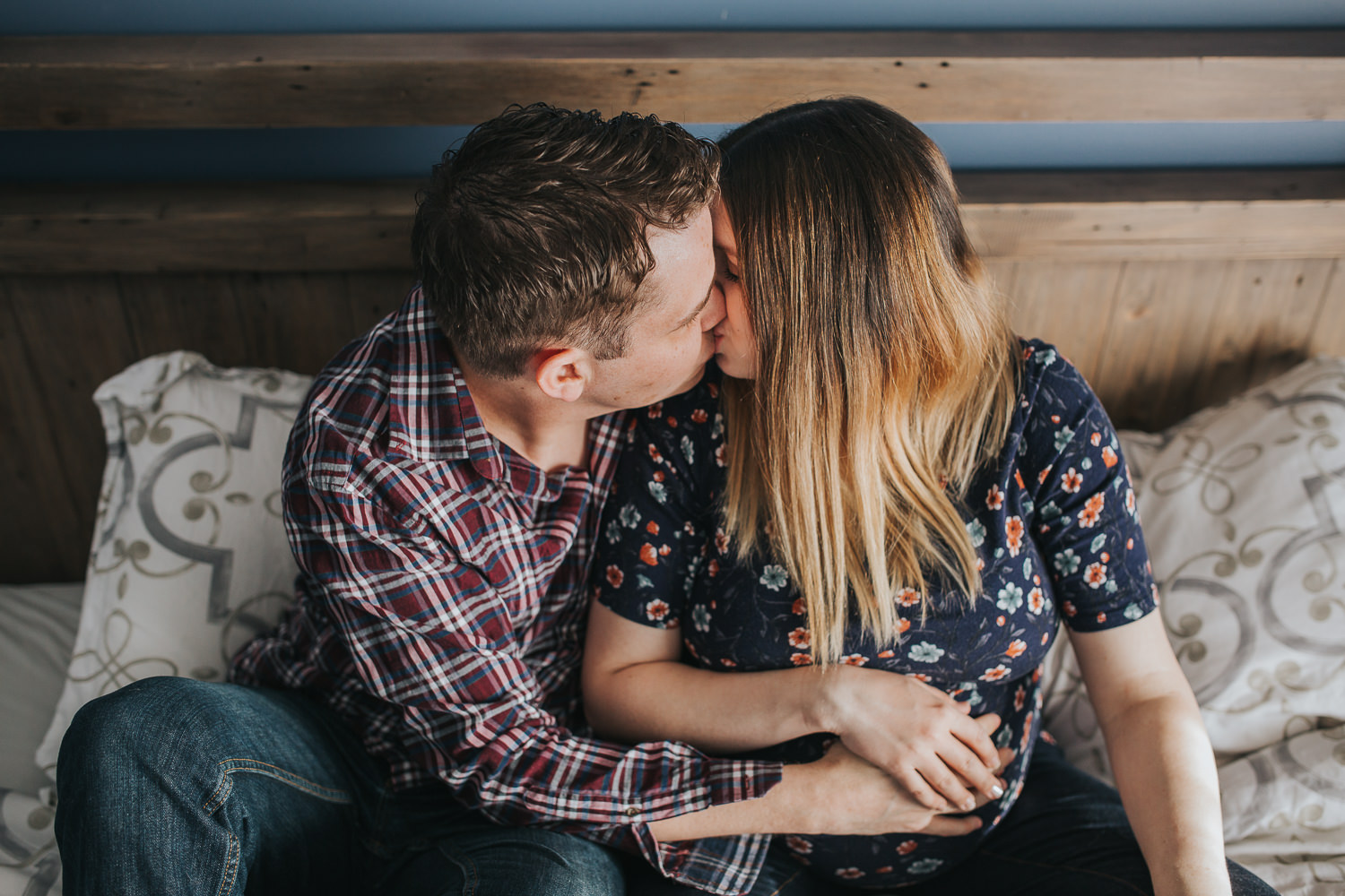 expecting couple kisses on bed, hands holding wife's pregnant belly - Markham In-Home Photography