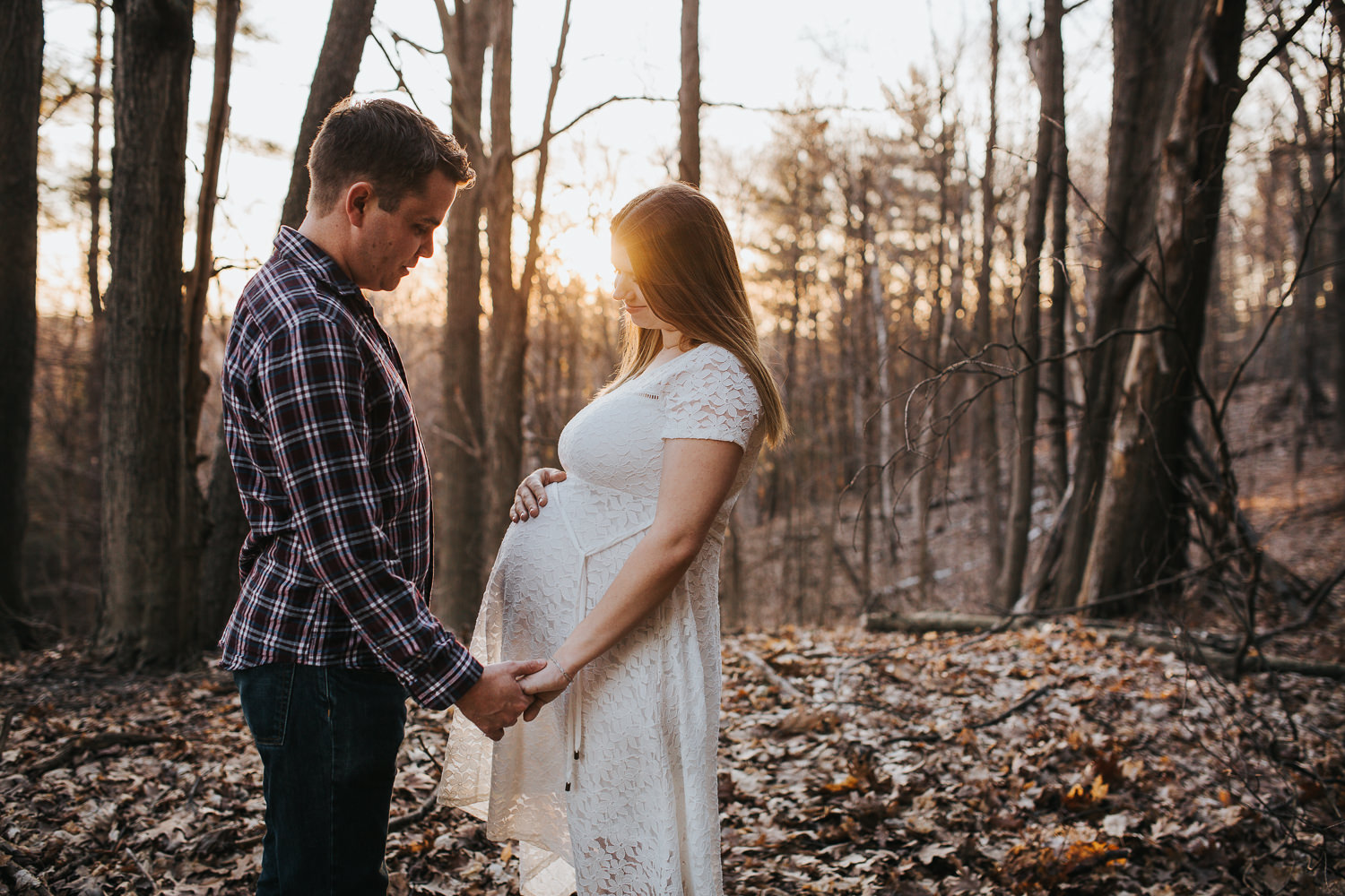 husband and pregnant wife stand in forest, looking at baby belly as the sun sets behind them - Stouffville Lifestyle Photos