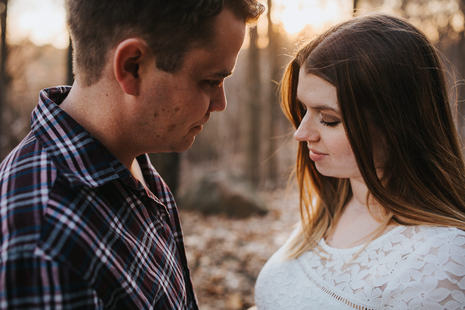 husband looks at pregnant wife as they stand in forest with the sun setting behind them - Markham Lifestyle Photos