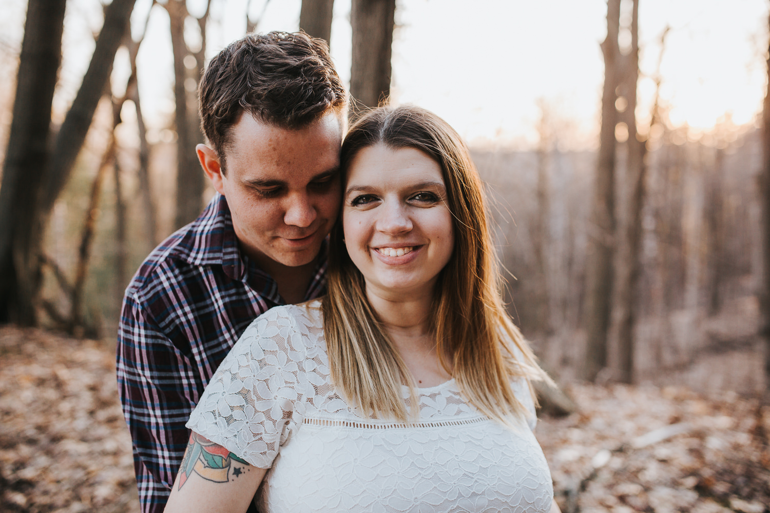 husband embraces pregnant wife, standing in forest with sunset behind them - Barrie Lifestyle Photos