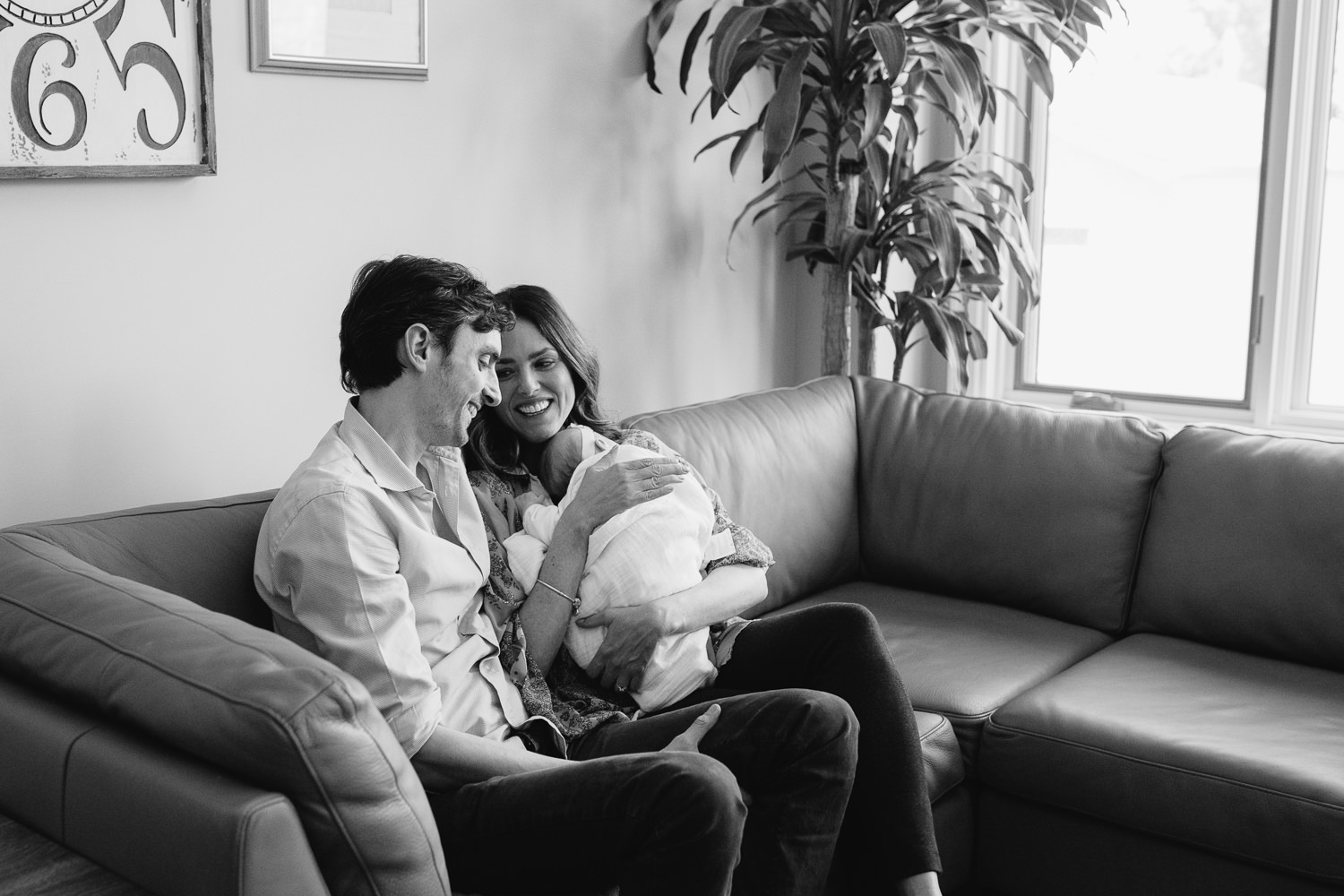 new parents sitting on couch holding 2 month old baby daughter, laughing - Newmarket In-Home Photography