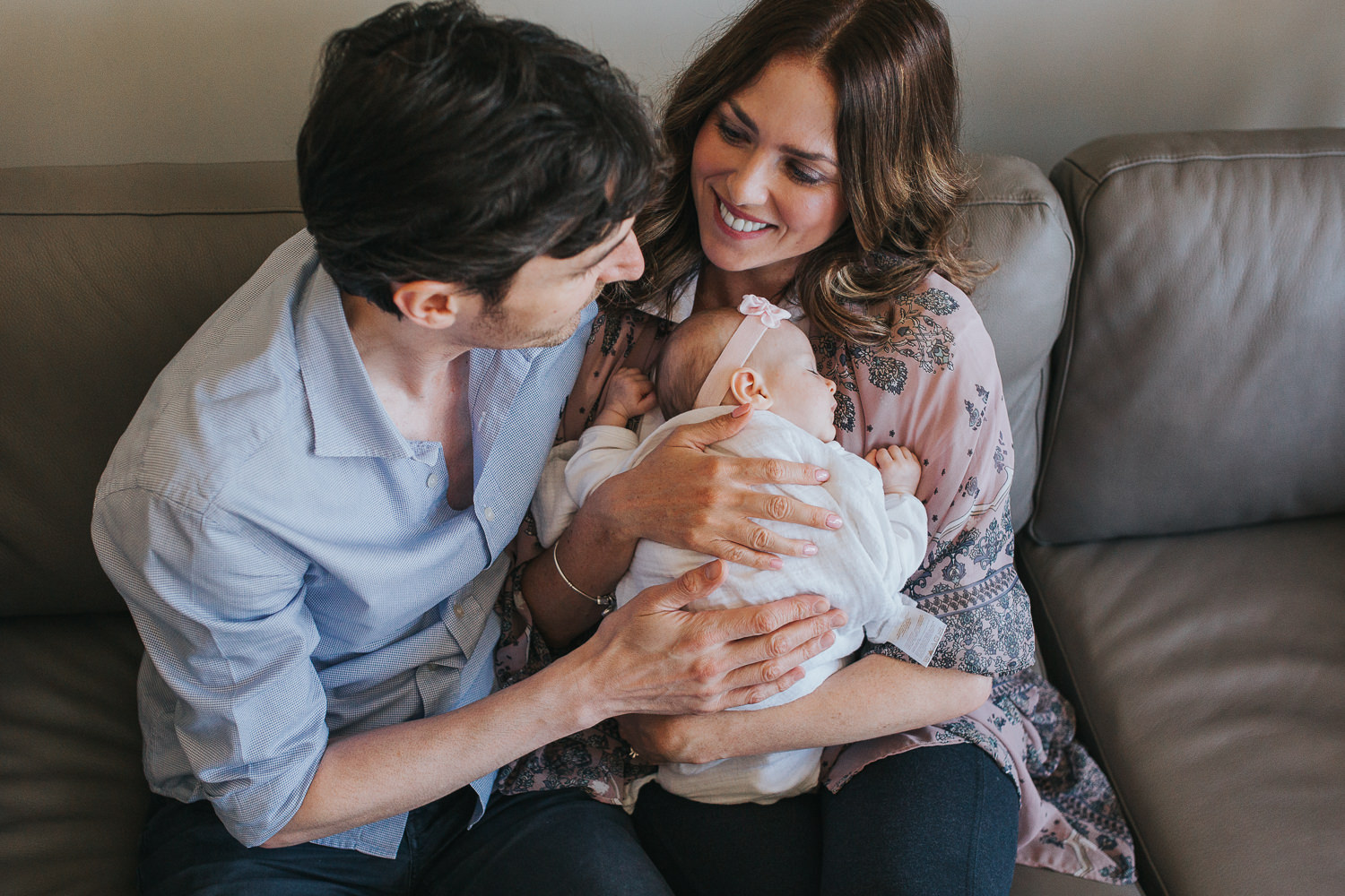 new parents sitting on couch holding 2 month old baby daughter, laughing - Stouffville In-Home Photography