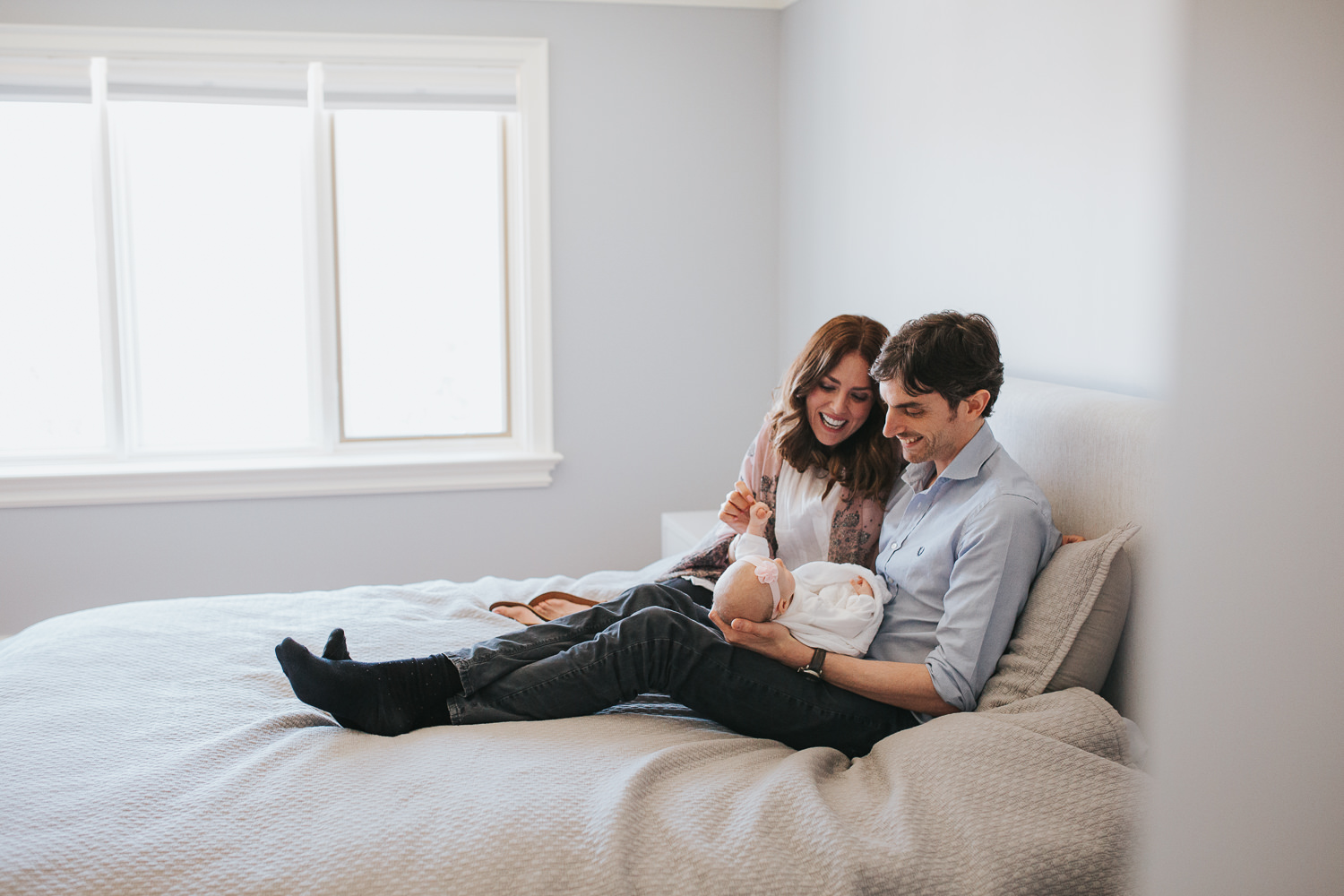 husband and wife sitting on master bed, dad holding 2 month old baby girl - Newmarket Lifestyle Photos