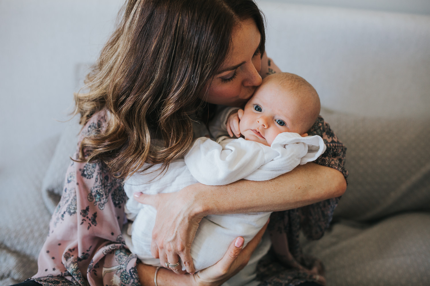 new mother sitting on bed kissing 2 month old baby girl, who is looking at camera - Markham In-Home Photos