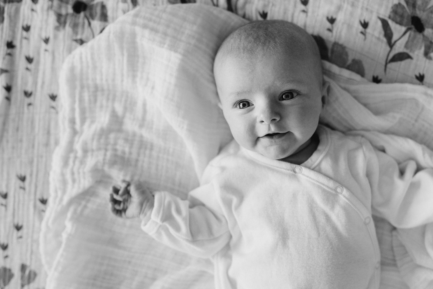 2 month old baby girl in white sleeper lying on bed looking at camera - Newmarket Lifestyle Photography