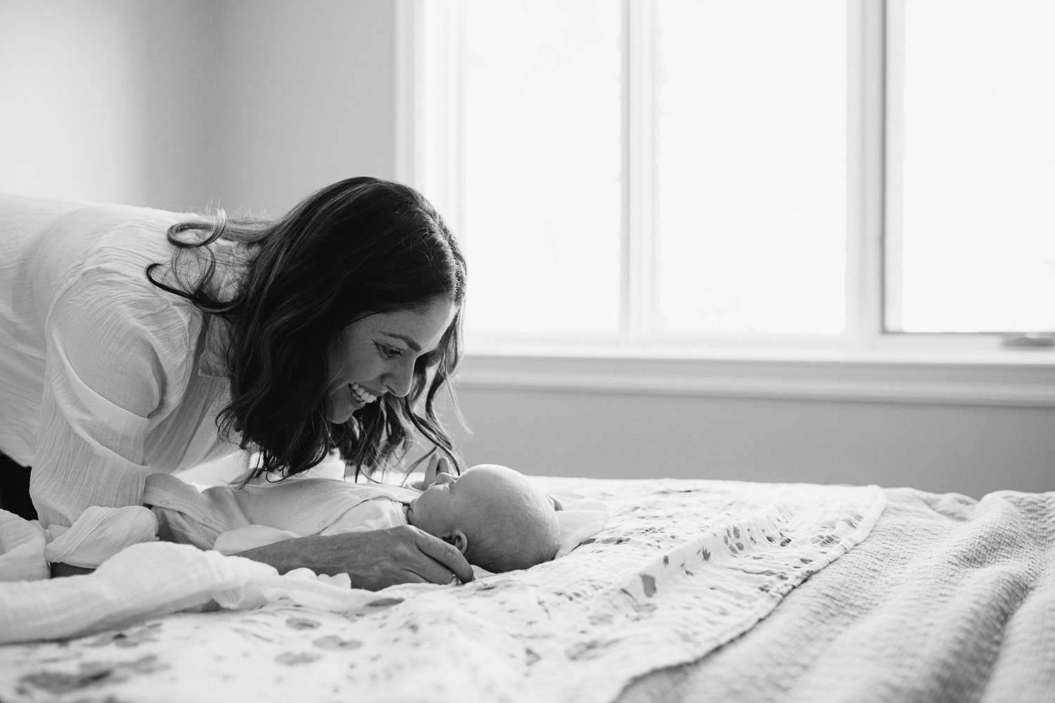 2 month old baby girl lying on bed, mom leaning over smiling at daughter - Barrie In-Home Photos