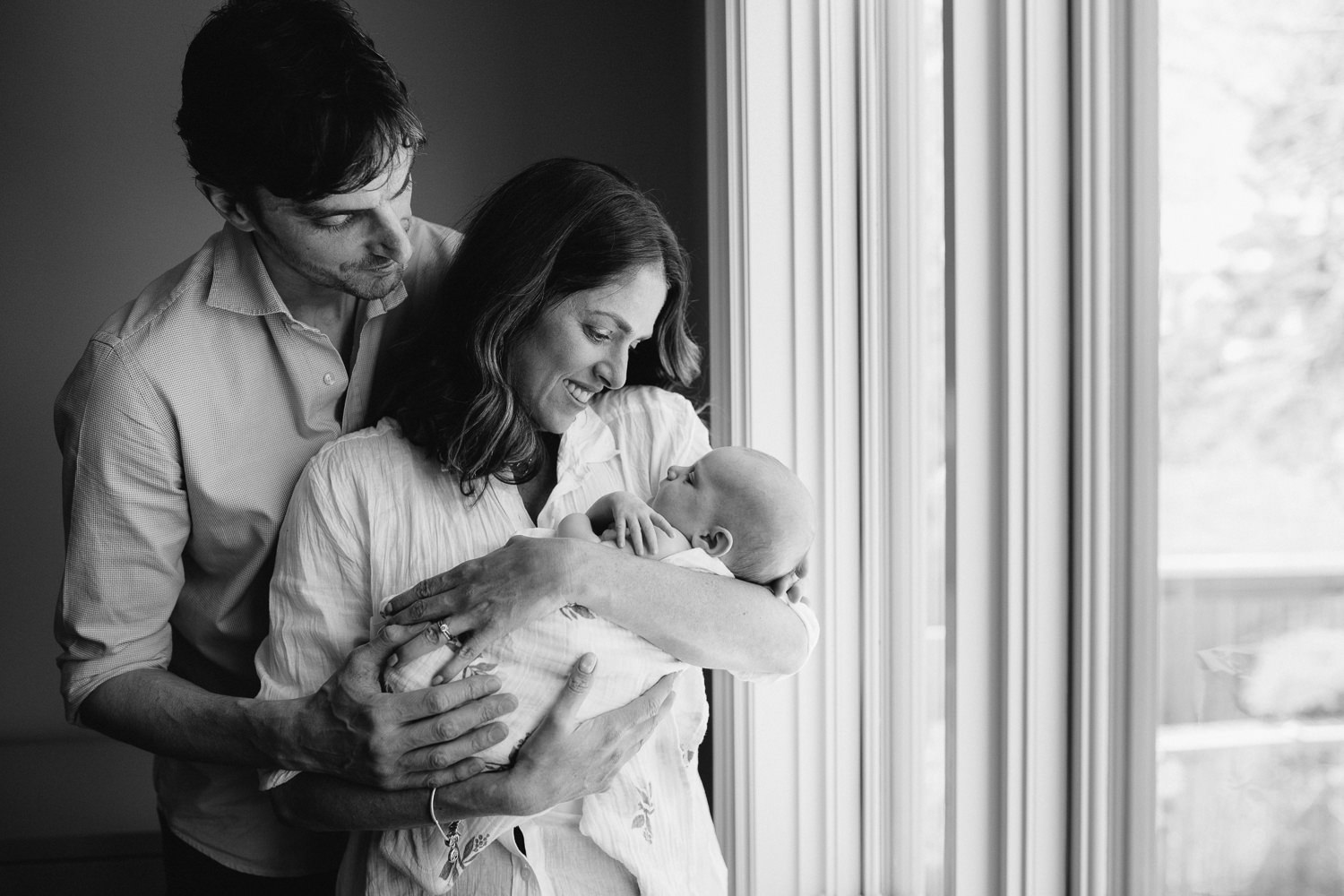 new parents stand at window, mom holding 2 month old daughter, dad embraces mom - Stouffville In-Home Photography
