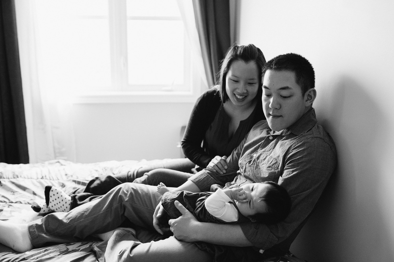 new parents sitting on master bed holding and looking at 3 month old baby girl - Stouffville In-Home Photos