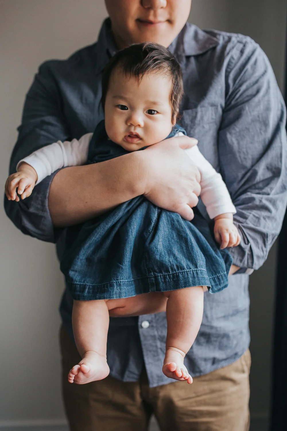 3 month old baby girl in blue dress sits in dad's arms, looking at camera - Barrie In-Home Photos