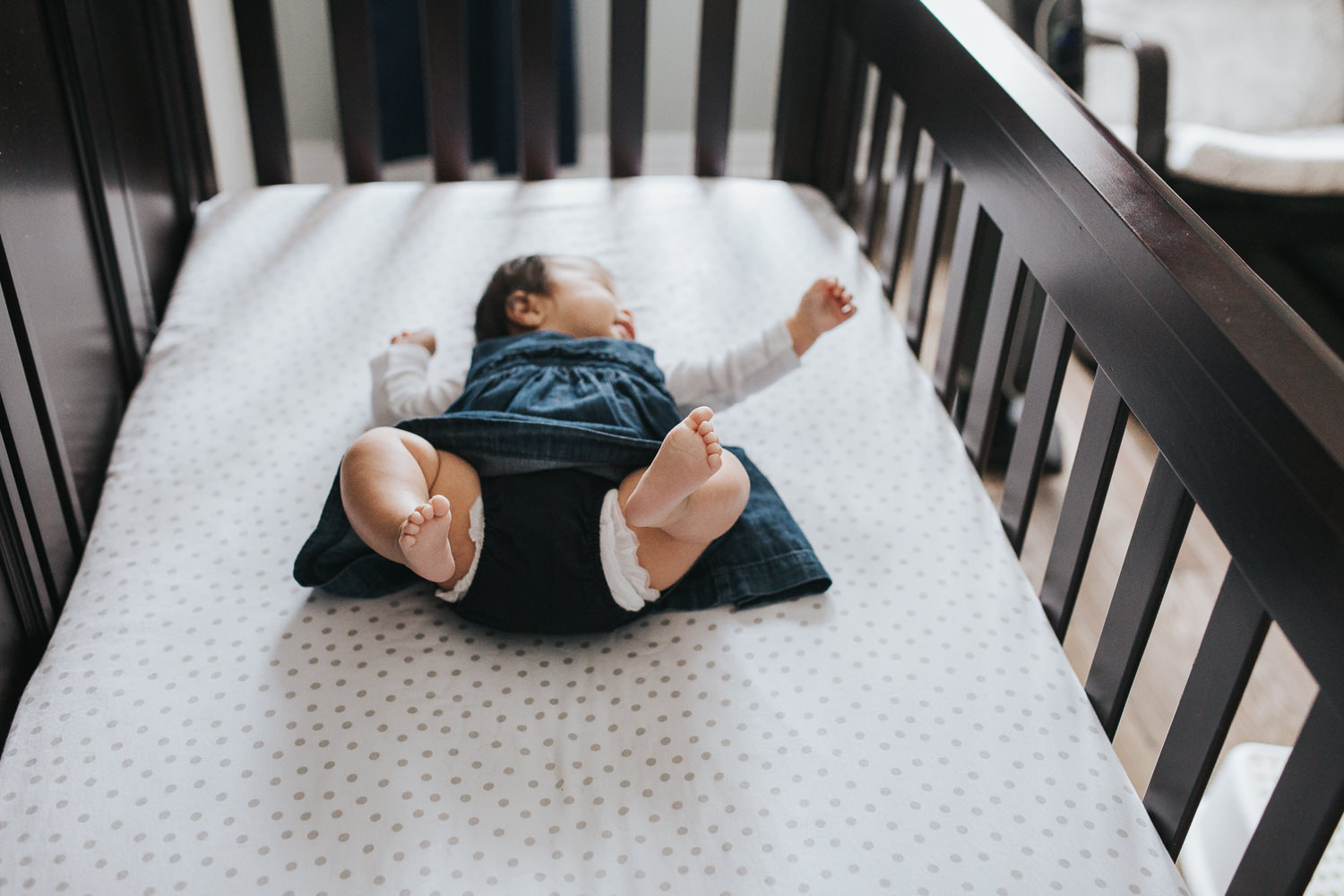 3 month old baby girl in blue dress lying in nursery crib with feet in air - Uxbridge Lifestyle Photography