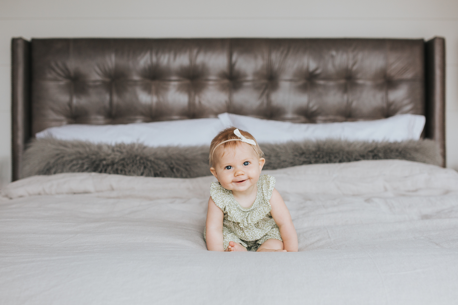 6 month old baby girl with blonde hair and blue eyes sitting on bed smiling at camera - Barrie In-home Family Photography