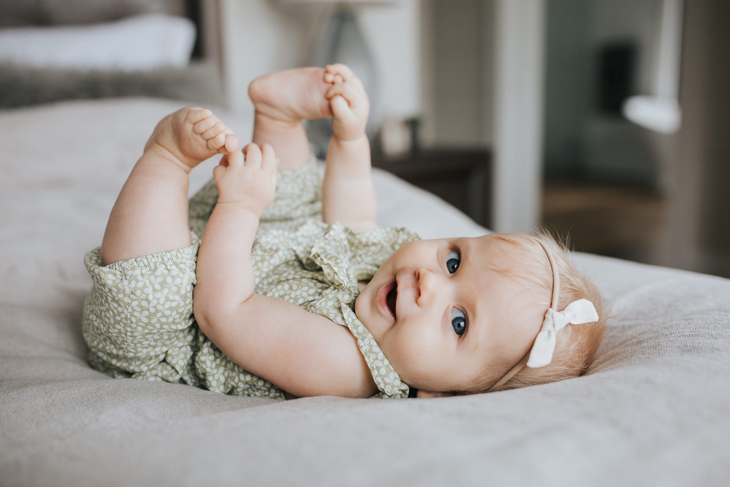 6 month old baby girl with blonde hair and blue eyes lying on bed playing with feet and looking at camera - Markham In-home Family Photography