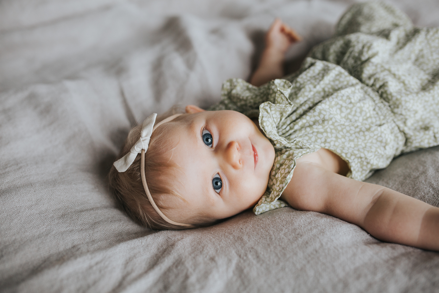 6 month old baby girl with blonde hair and blue eyes lying on bed looking at camera - Markham In-home Family Photos