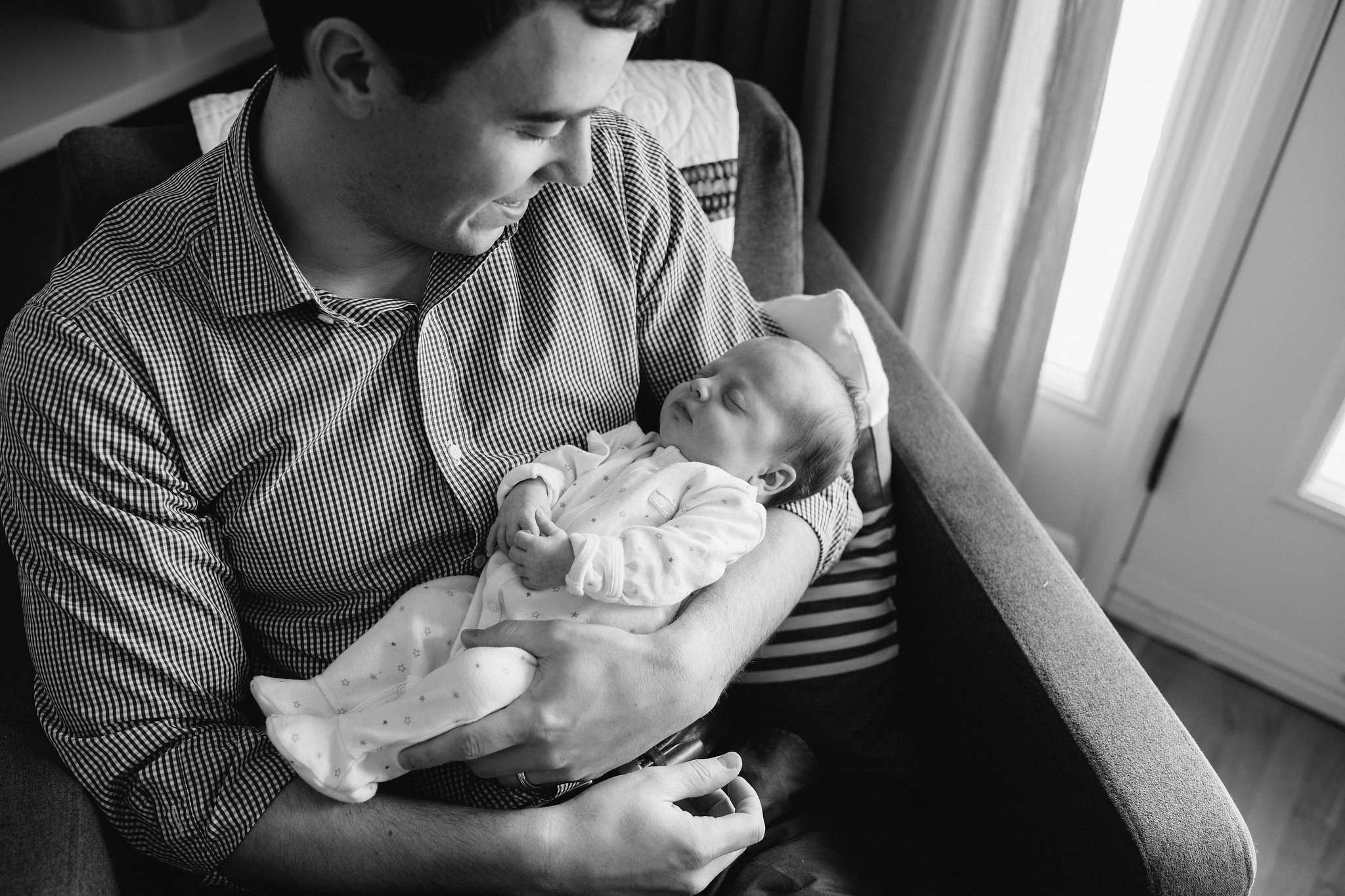 first time dad sitting in chair holding 4 week old baby girl in sleeper with her hands folded - Barrie Lifestyle Photography