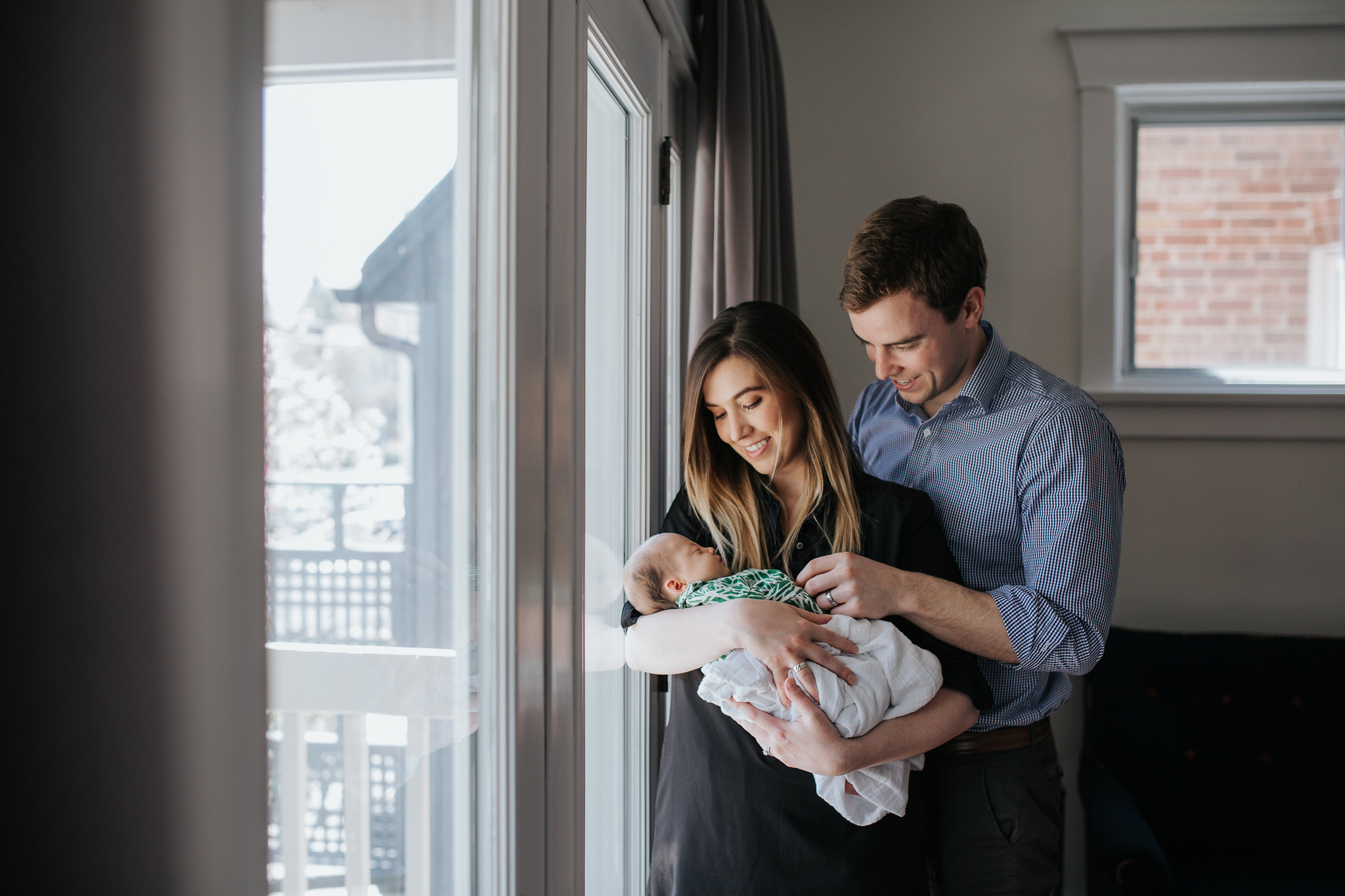 new parents standing at window holding and looking at 4 week old baby girl in swaddle - Barrie Lifestyle Photos