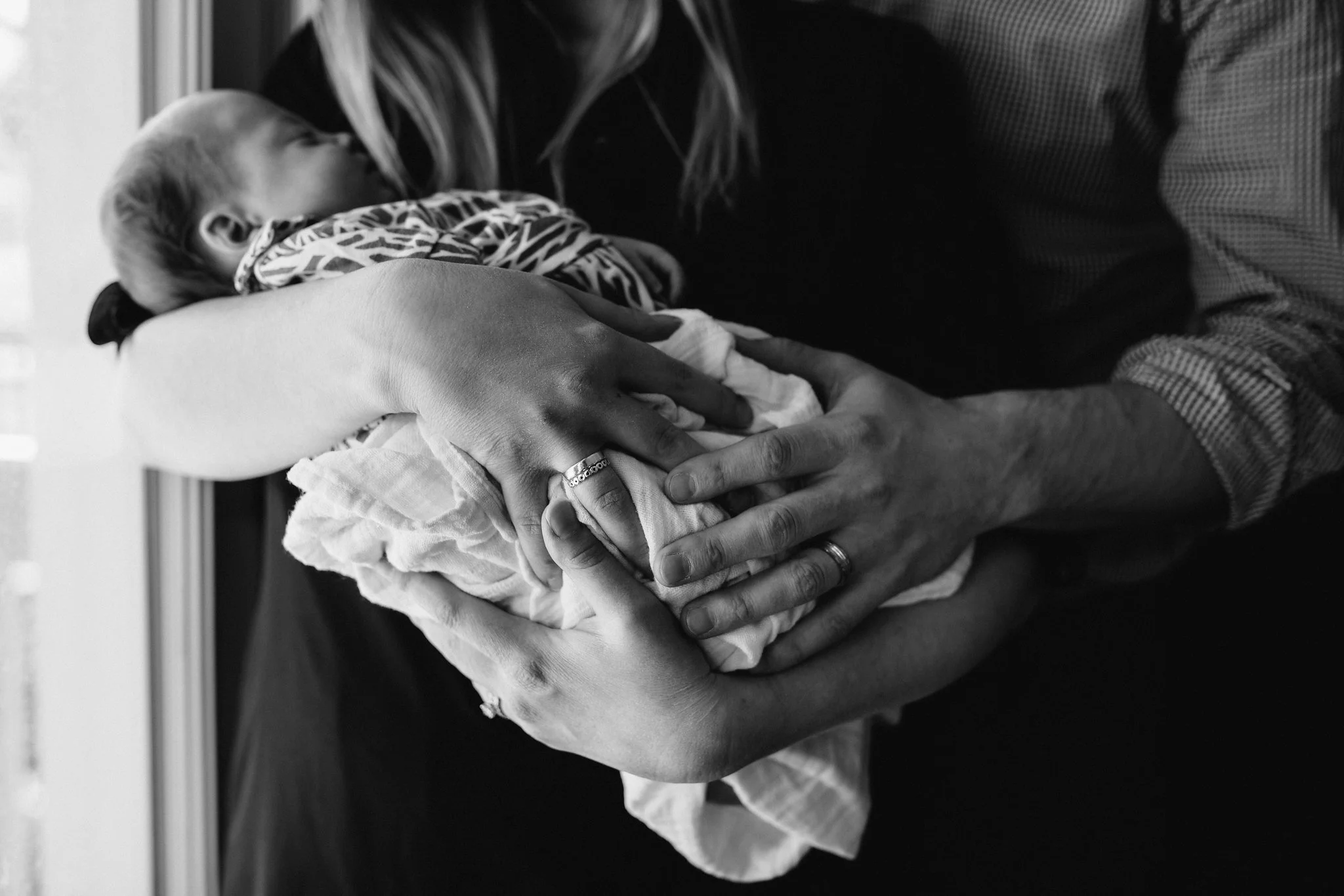 new parents standing at window holding 4 week old baby girl in swaddle - Newmarket Lifestyle Photography