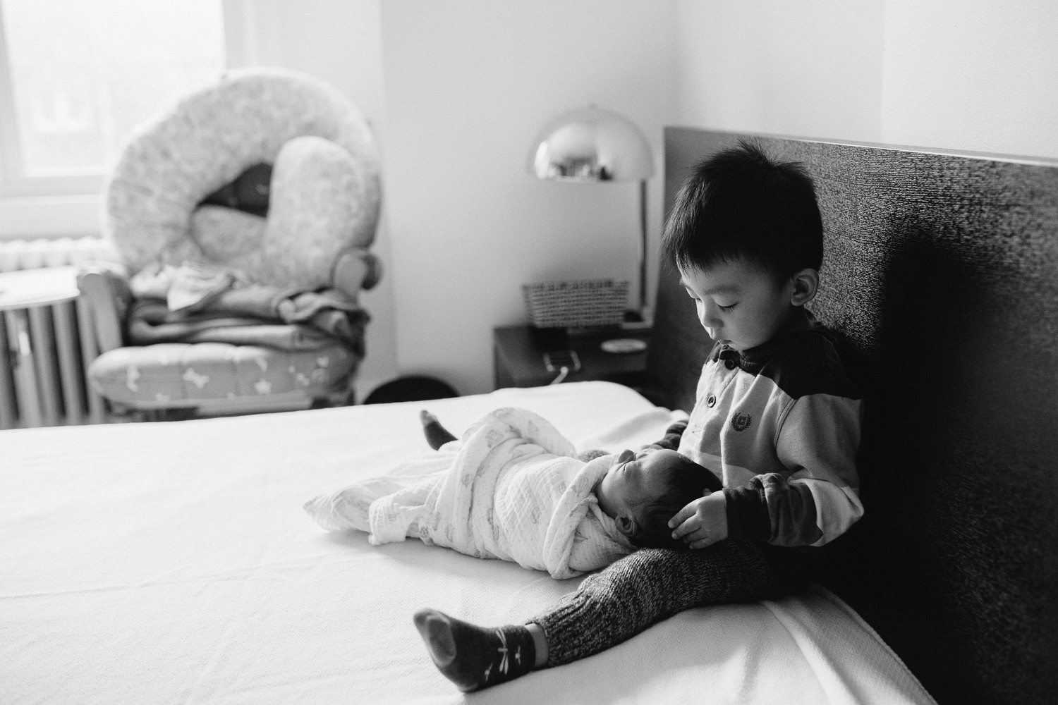 2 year old toddler boy sitting on bed holding 2 week old baby brother - Barrie In-Home Photography