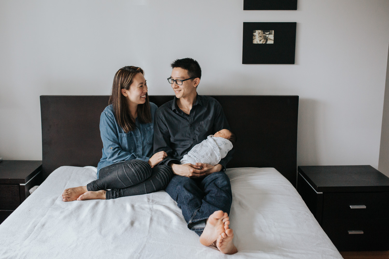 husband and wife sitting on bed, husband holding 2 week old baby boy, wife smiling at husband - Barrie In-Home Photos