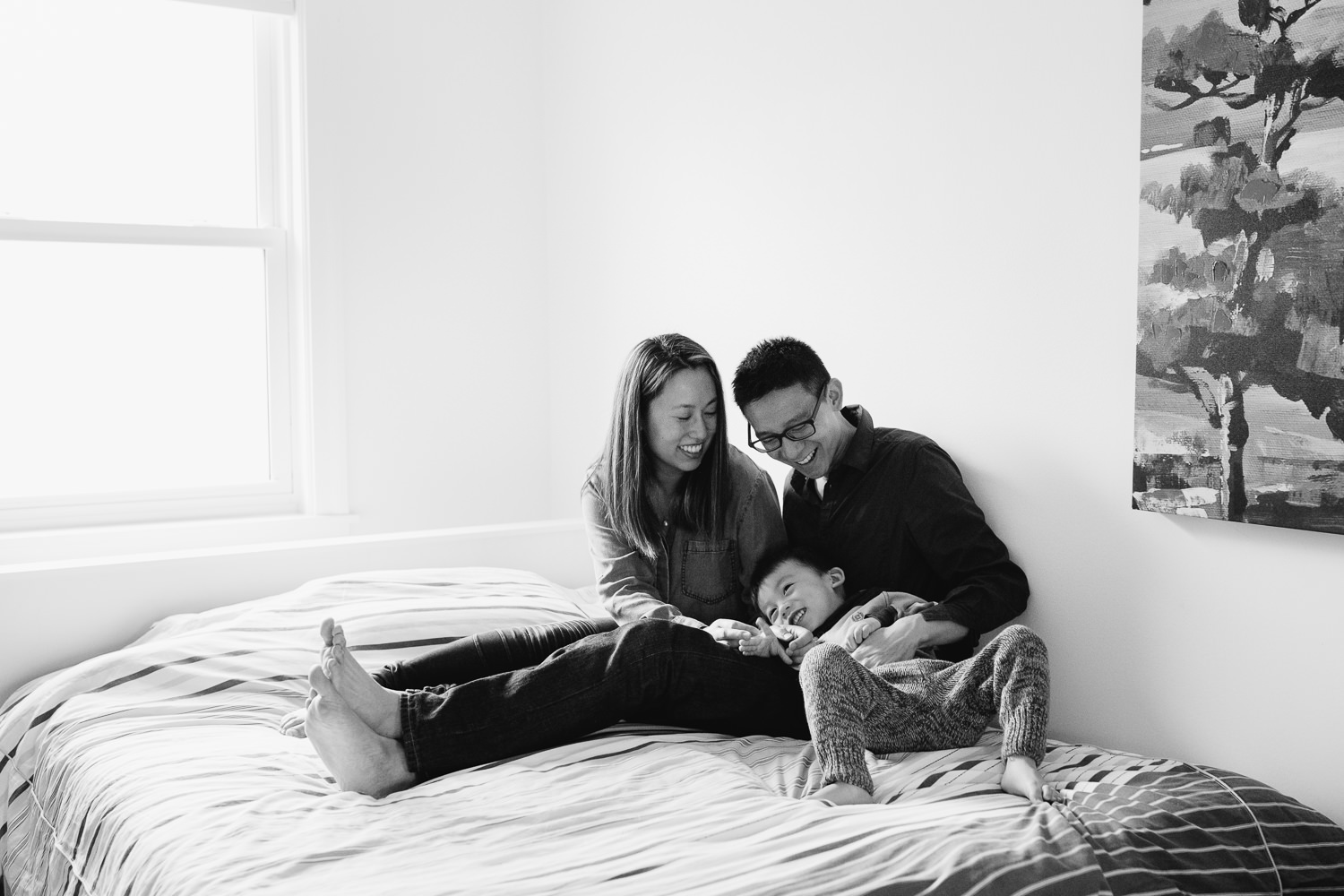 mom and dad sitting on bed ticking laughing 2 year old toddler boy - Newmarket Lifestyle Photos