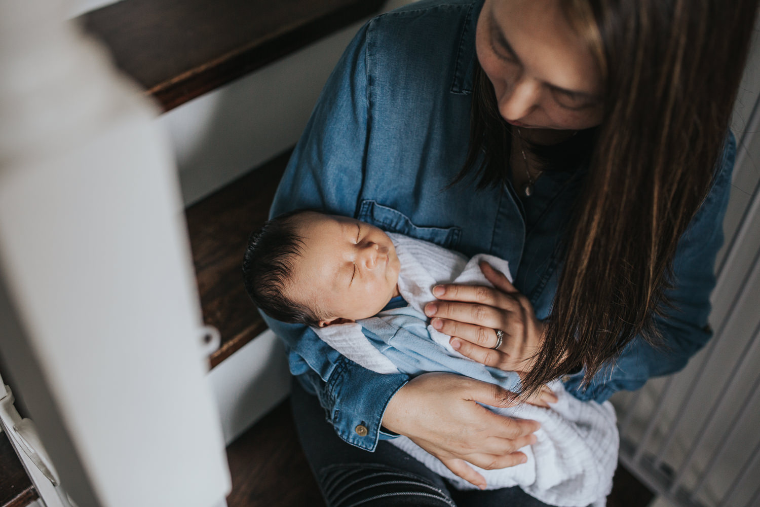 mom in blue shirt with dark hair sits on stairs holding 2 week old baby boy - Barrie Lifestyle Photography