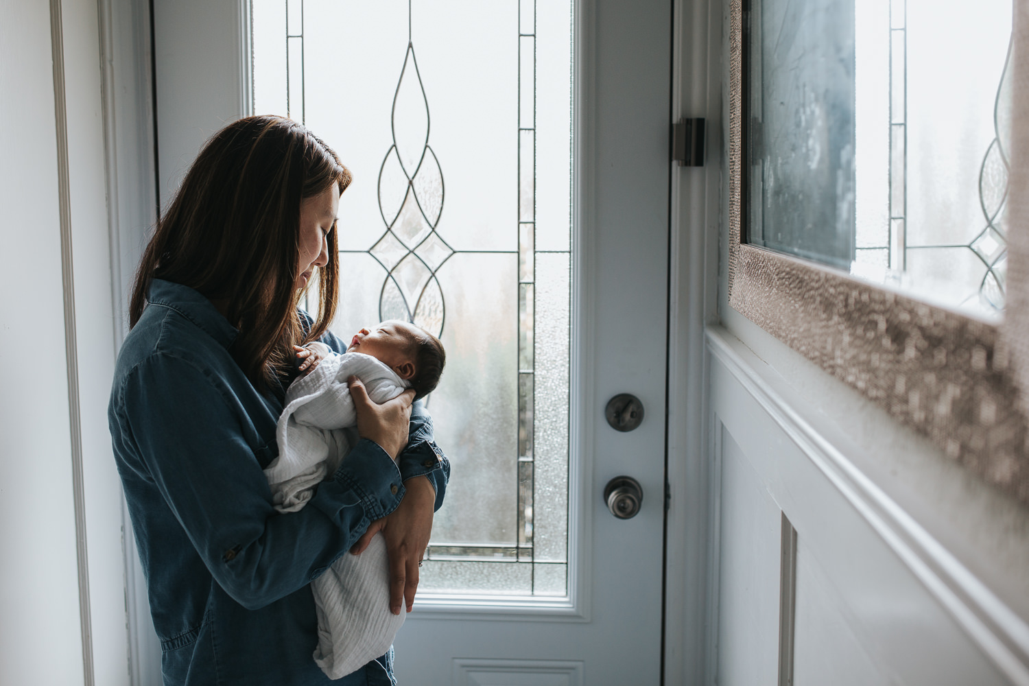 mom in blue shirt with dark hair stands at door holding 2 week old baby boy - Barrie In-Home Photography