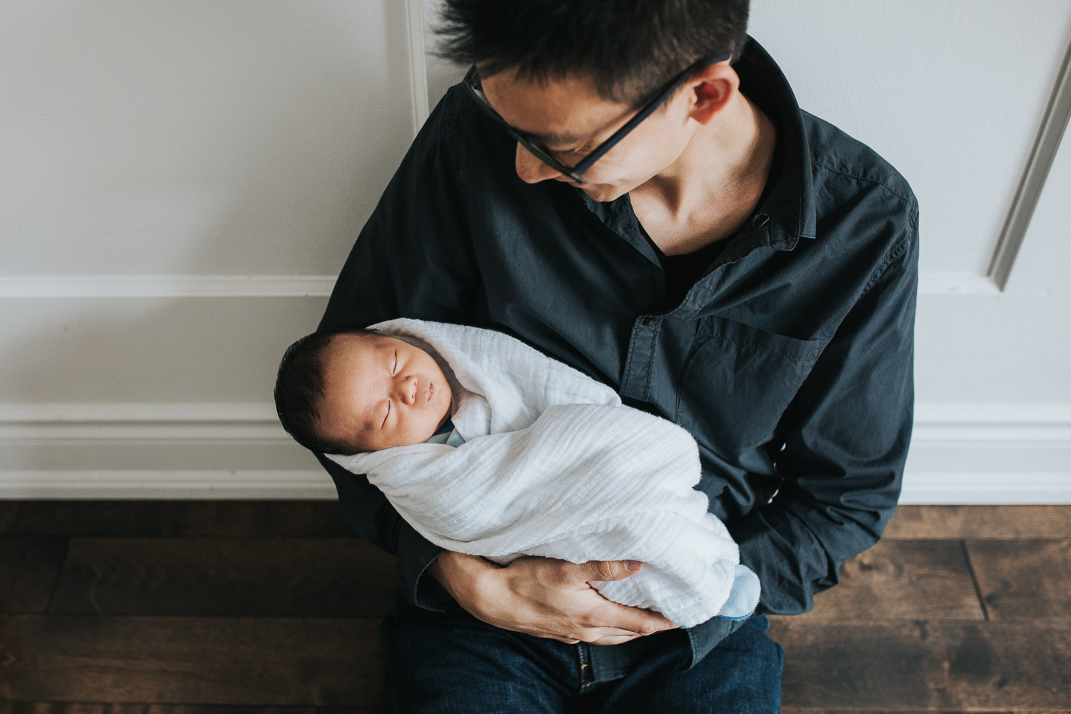 father sitting on floor holding 2 week old baby boy sleeping in white swaddle - Markham In-Home Photos