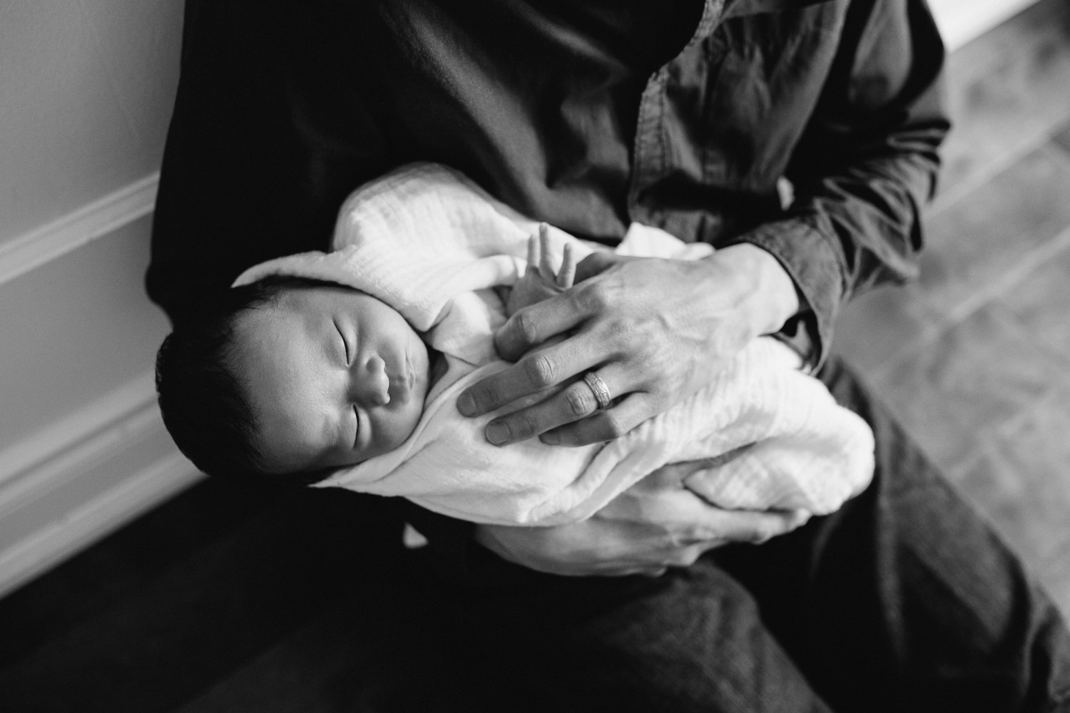 father sitting on floor holding 2 week old baby boy sleeping in white swaddle - Barrie In-Home Photos