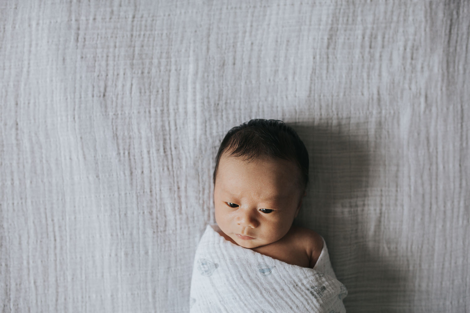 2 week old baby boy with eyes open lying on bed in blue and white swaddle - Newmarket Lifestyle Photography
