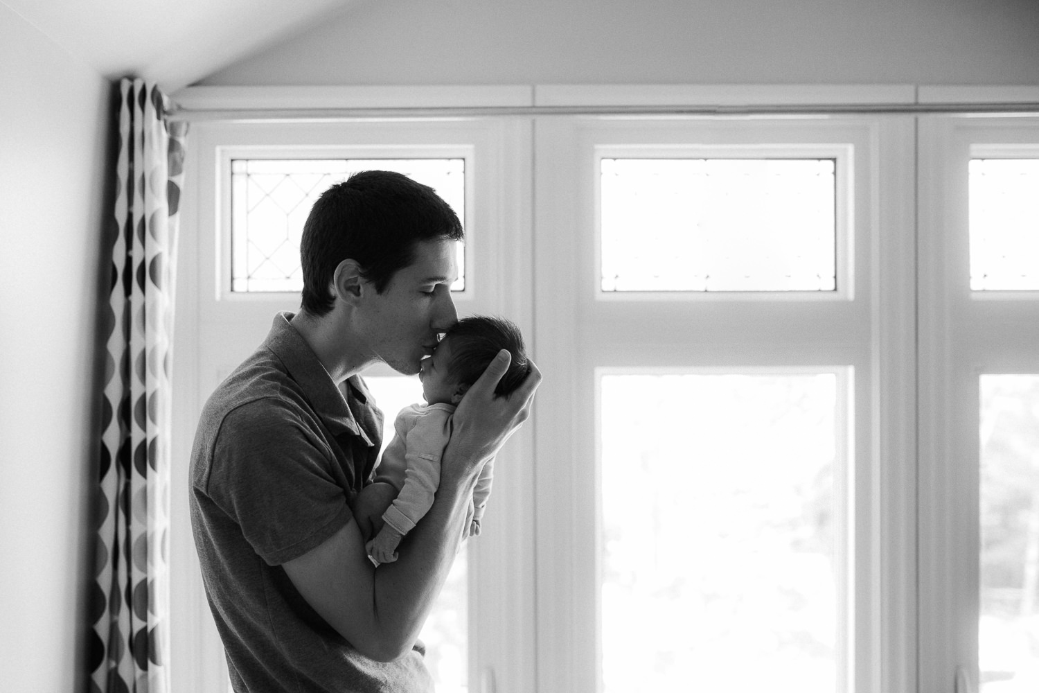 first time dad holding 2 week old baby girl in front of window, kissing her face - Stouffville In-Home Photography