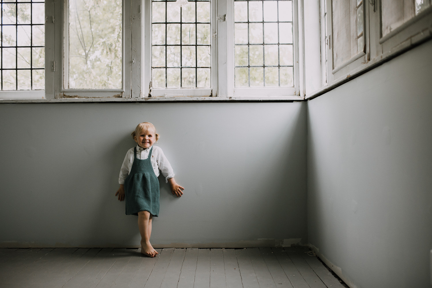 2 year old blonde toddler girl in blue dress stands leaning against wall and smiling at camera - Stouffville In-Home Family Photography
