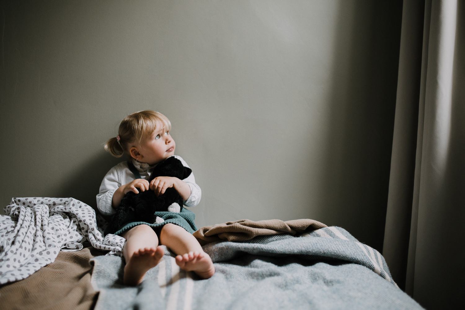 2 year old blonde toddler girl sits on bed holding stuffed cat and looking out bedroom window - Barrie In-Home Family Photography
