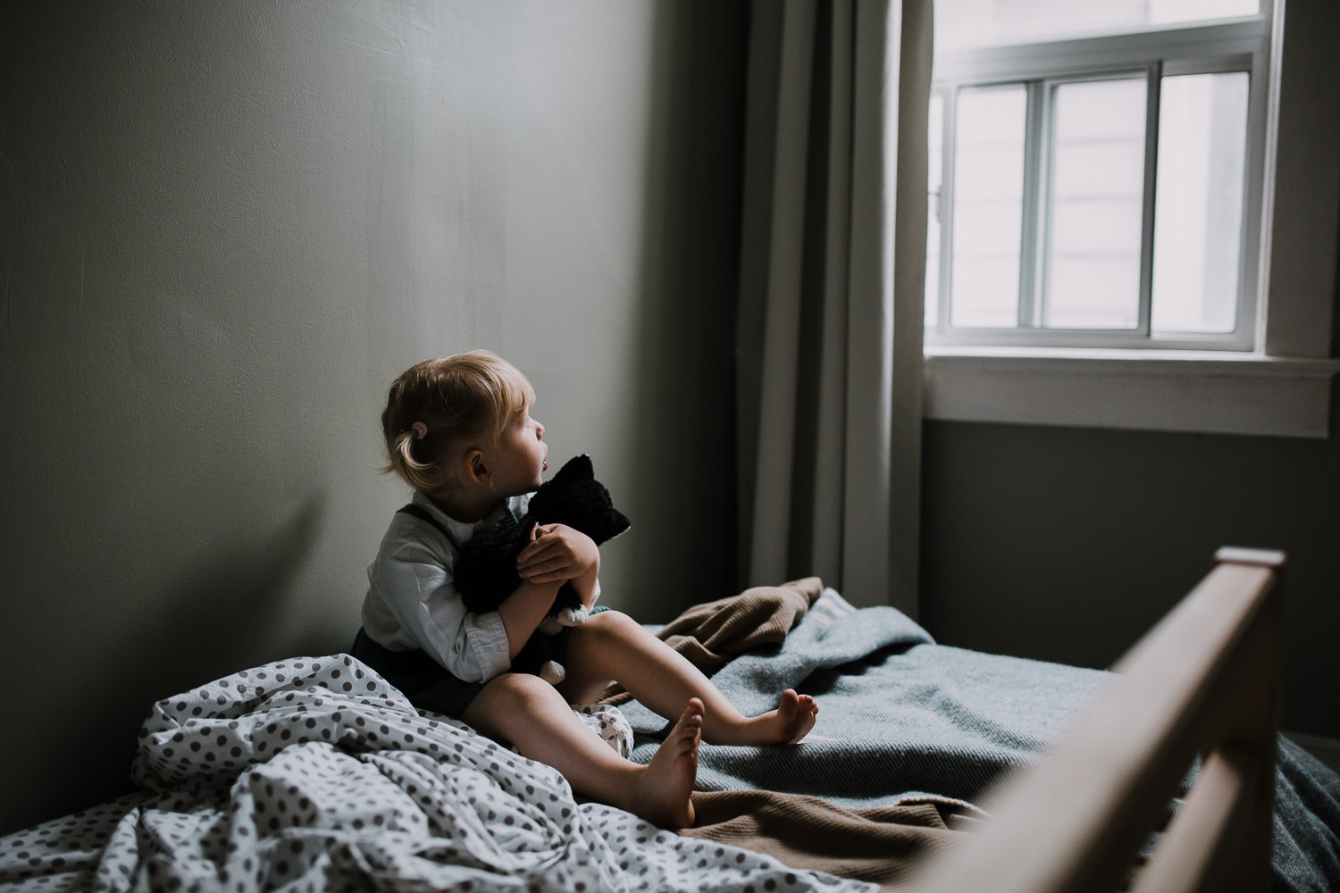 2 year old blonde toddler girl sits on bed holding stuffed cat and looking out bedroom window - Markham In-Home Family Photography