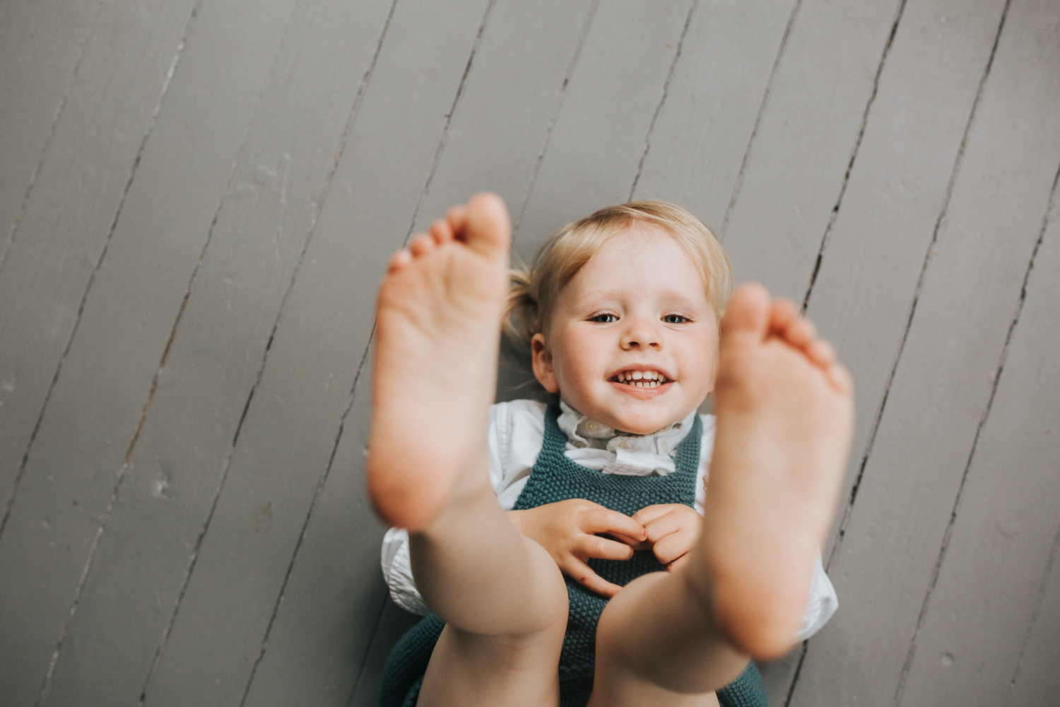 2 year old toddler girl with blonde hair and blue eyes lying on floor looking at camera with feet in the air - Newmarket In-Home Family Photography