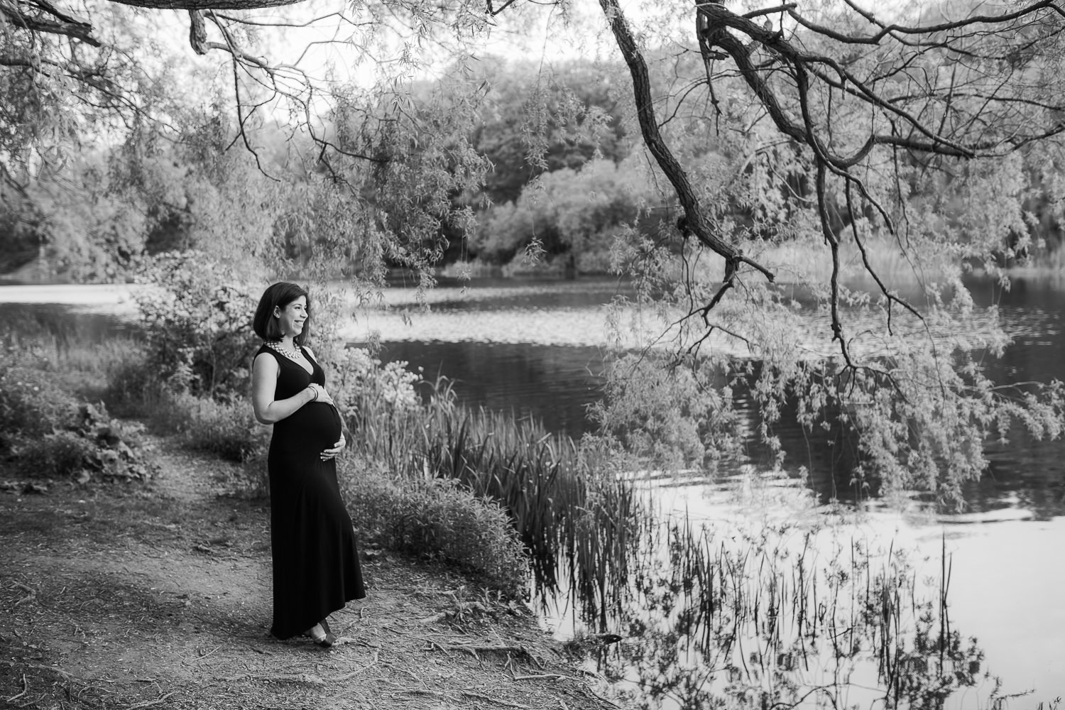 pregnant brunette woman in black dress stands beside river holding baby bump - Stouffville Lifestyle Photography