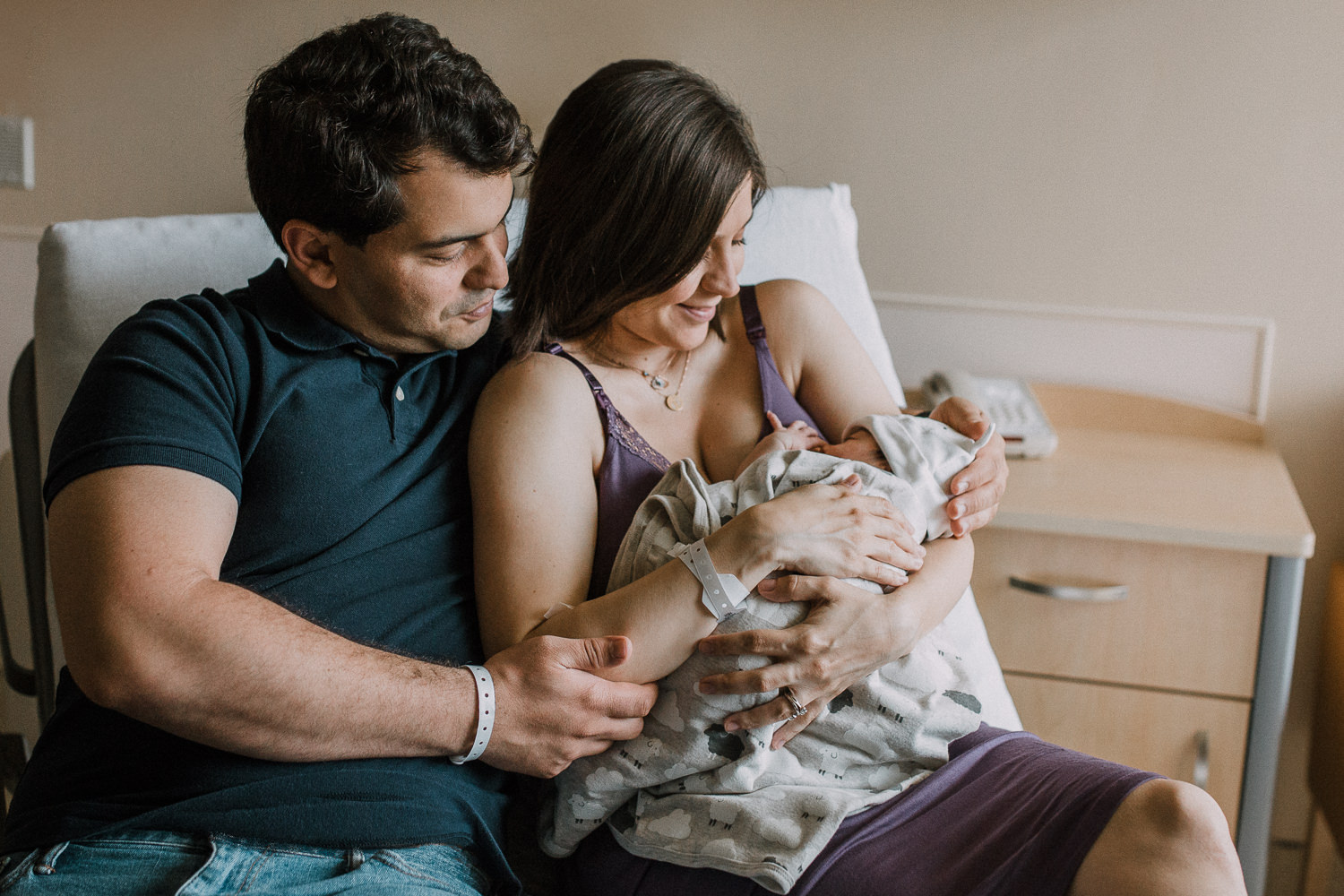 brand new family of three sits on hospital bed, mom holding 1 day old baby girl - Newmarket In-Hospital Photography