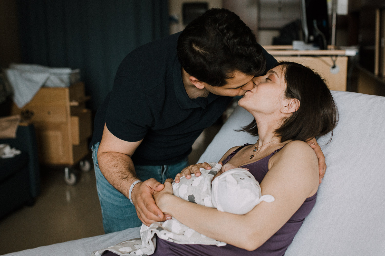 new mom sits on hospital bed holding 1 day old baby girl as dad leans over and kisses wife - Markham In-Hospital Photography