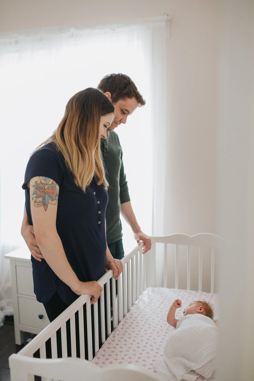 parents stand in nursery looking at 2 week old swaddled baby girl with red hair asleep crib - Newmarket Lifestyle Photos