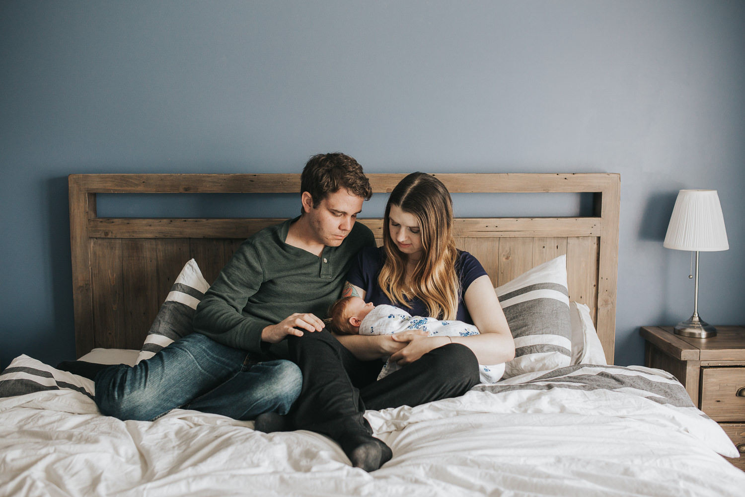 family of 3, new parents sitting on bed holding and looking at 2 week old baby girl with red hair - Newmarket Lifestyle Photography